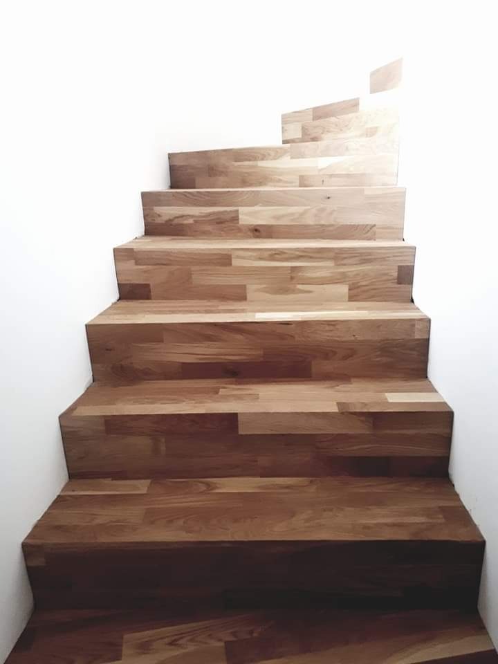 Drewniane schody z parkietu ułożonego w cegiełkę, prowadzące w górę wzdłuż białej ściany, oświetlone od góry.