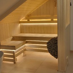 MAS-KOMINKI Krzysztof Sobczyk - Jasne, drewniane wnętrze sauny z oświetleniem LED pod ławkami i kamieniami w koszu, umieszczone na poddaszu ze skośnym sufitem.