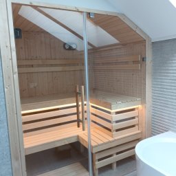 MAS-KOMINKI Krzysztof Sobczyk - Wnętrze sauny z drewna z przeszklonymi drzwiami, oświetleniem LED pod ławkami i widocznym piecem z boku.