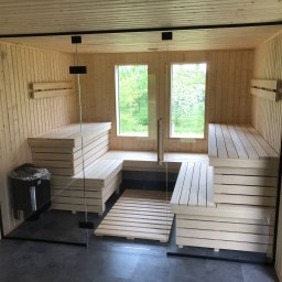 MAS-KOMINKI Krzysztof Sobczyk - Wnętrze nowej sauny z jasnego drewna, z przeszklonym wejściem i widokiem na zieleń przez dwa okna, z piecem elektrycznym w rogu i drewnianym rusztem na podłodze.