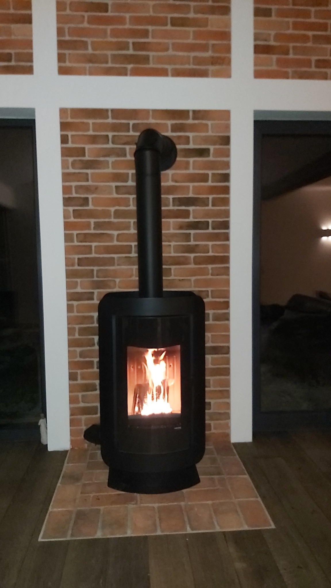 Czarny piec wolnostojący z płomieniami, ustawiony na ceglanej podłodze przed ścianą z cegły, z widocznymi białymi ramami okiennymi po bokach.