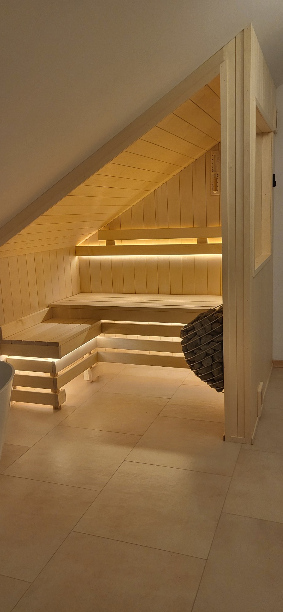 Jasne, drewniane wnętrze sauny z oświetleniem LED pod ławkami i kamieniami w koszu, umieszczone na poddaszu ze skośnym sufitem.