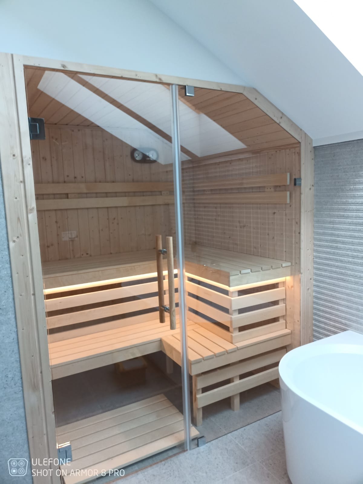Wnętrze sauny z drewna z przeszklonymi drzwiami, oświetleniem LED pod ławkami i widocznym piecem z boku.