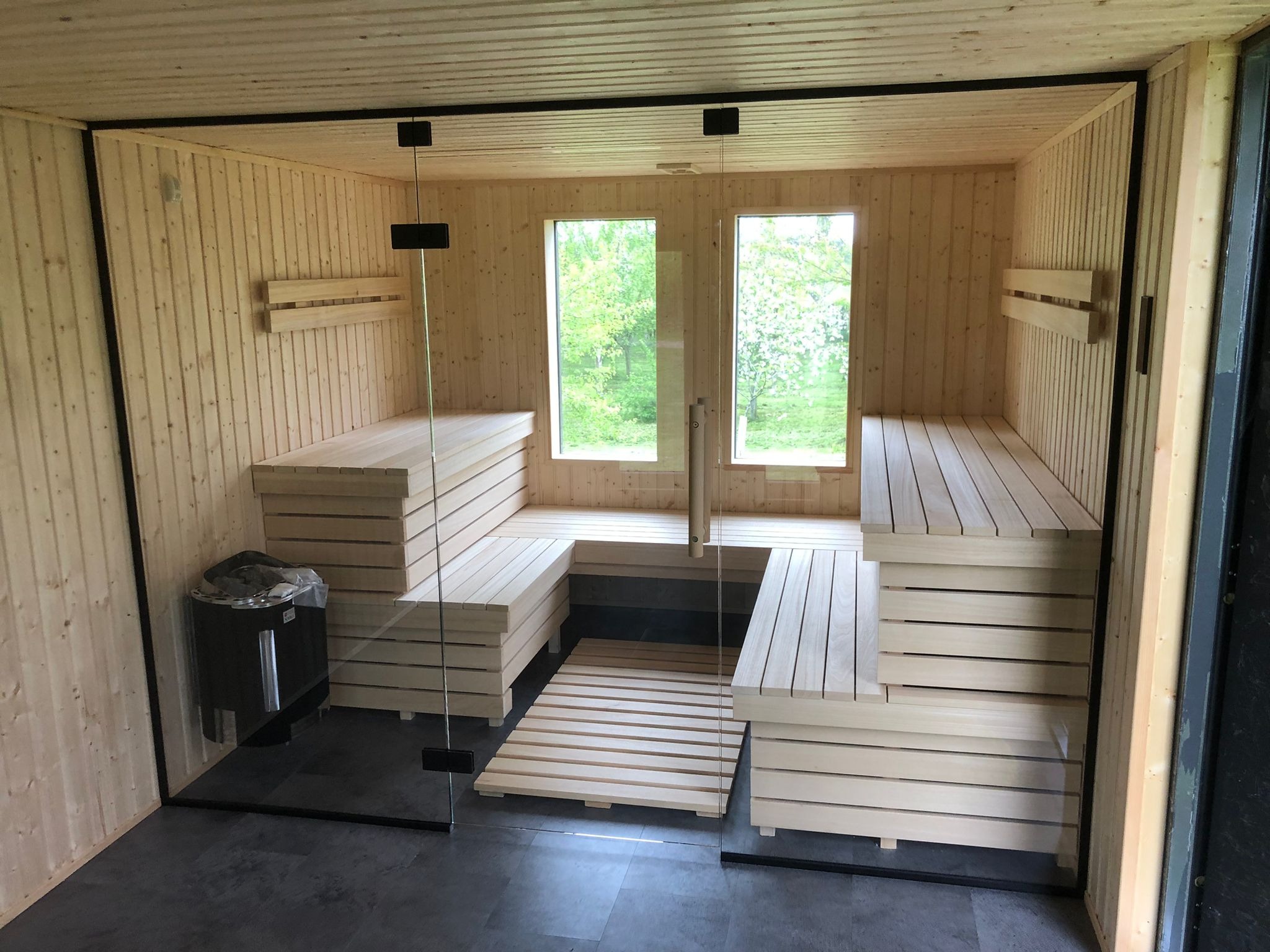 Wnętrze nowej sauny z jasnego drewna, z przeszklonym wejściem i widokiem na zieleń przez dwa okna, z piecem elektrycznym w rogu i drewnianym rusztem na podłodze.
