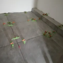 Szare płytki podłogowe w trakcie układania, z użyciem zielonych i czerwonych klinów poziomujących, widoczne fugi i narożnik pomieszczenia.