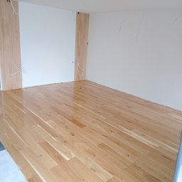Grzegorz Bujak - Jasne, lakierowane panele podłogowe w nowym, pustym pokoju z przygotowanymi przewodami elektrycznymi na ścianach.