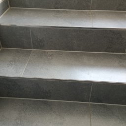 REMBUD Grzegorz Bujak - Szare, kamienne schody wyłożone płytkami z metalowymi listwami wykończeniowymi, prowadzące do góry, z doniczką widoczną w tle.