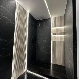 Nowoczesna łazienka z czarnymi marmurowymi ścianami, podświetlanymi panelami ściennymi i oświetleniem LED na suficie i podłodze, tworzącymi geometryczne wzory.