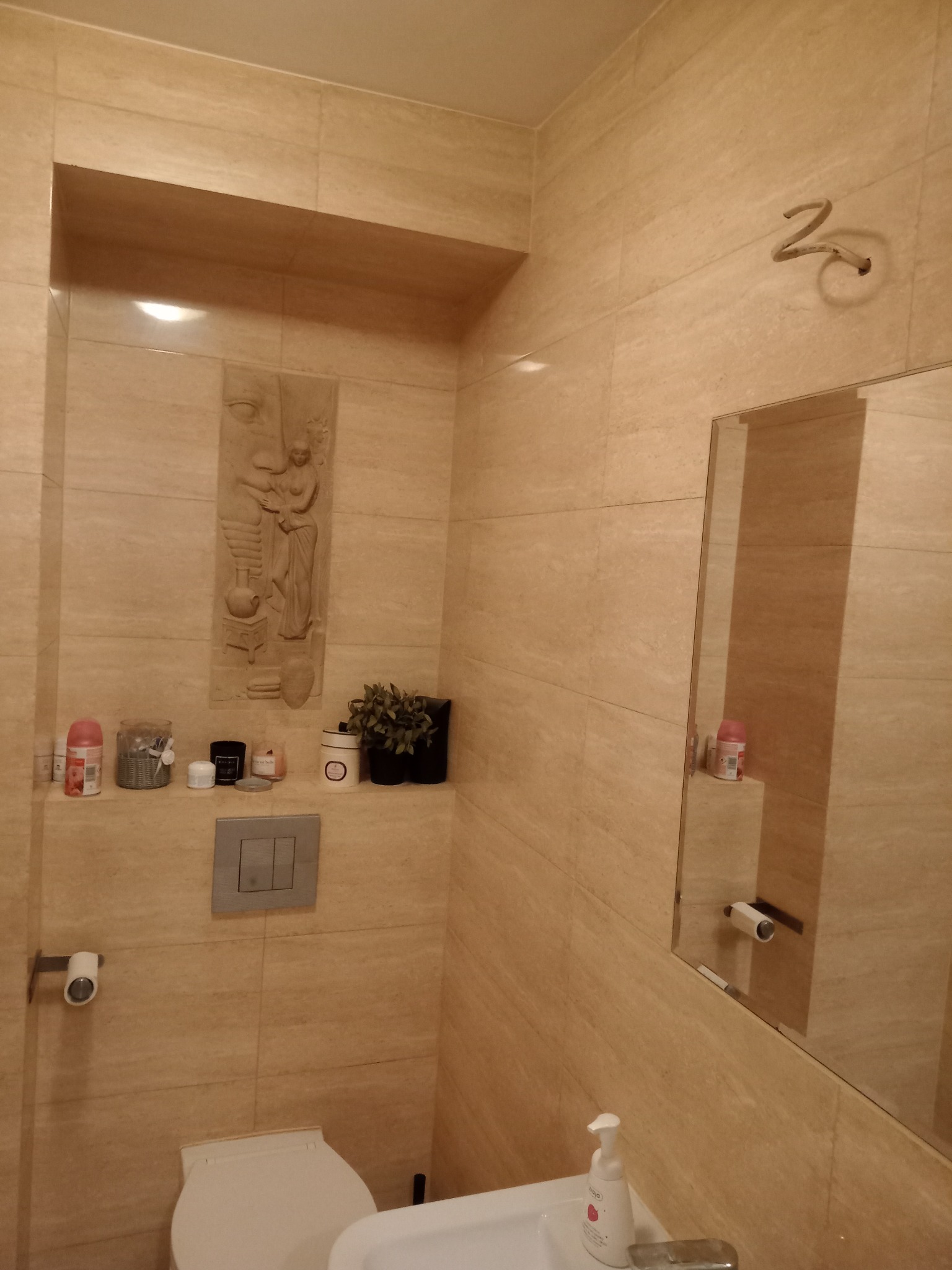 Beżowa łazienka z płytkami imitującymi trawertyn, z toaletą, umywalką, lustrem i półką z dekoracjami, wnęka z płaskorzeźbą.