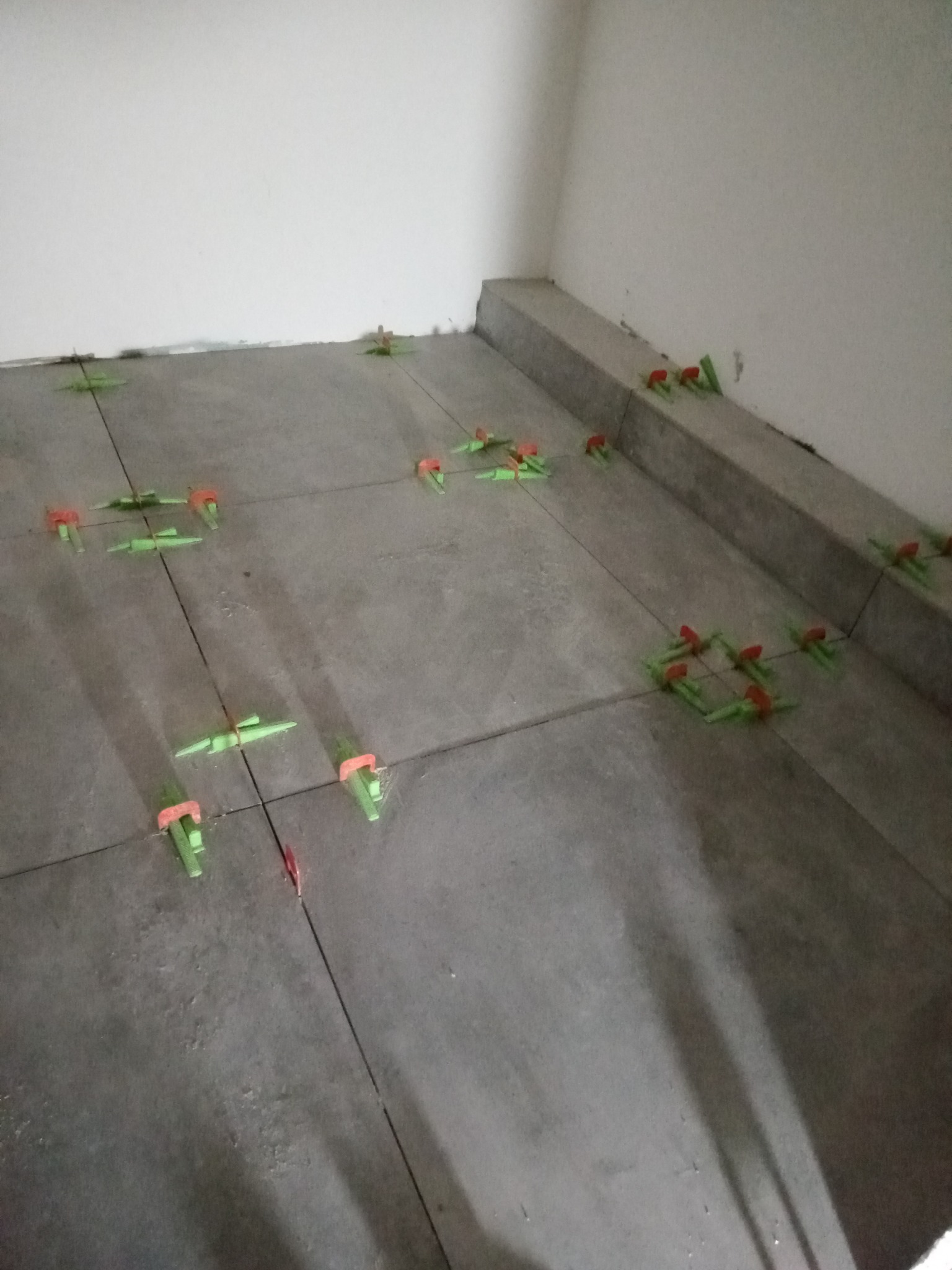 Szare płytki podłogowe w trakcie układania, z użyciem zielonych i czerwonych klinów poziomujących, widoczne fugi i narożnik pomieszczenia.