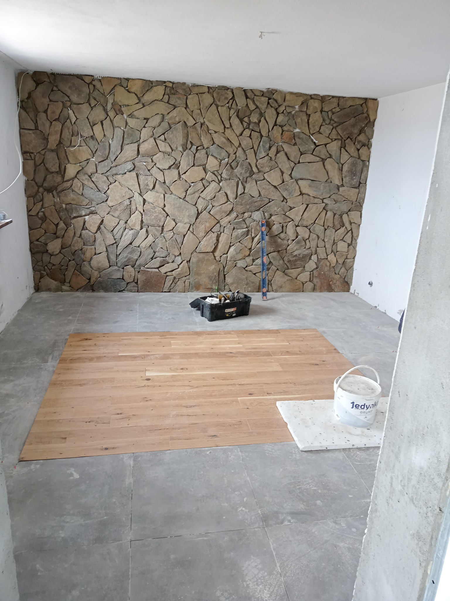 Fragment układania paneli podłogowych w pokoju z dekoracyjną ścianą z kamienia, widoczny kubeł z gruntem Jedynka i narzędzia.