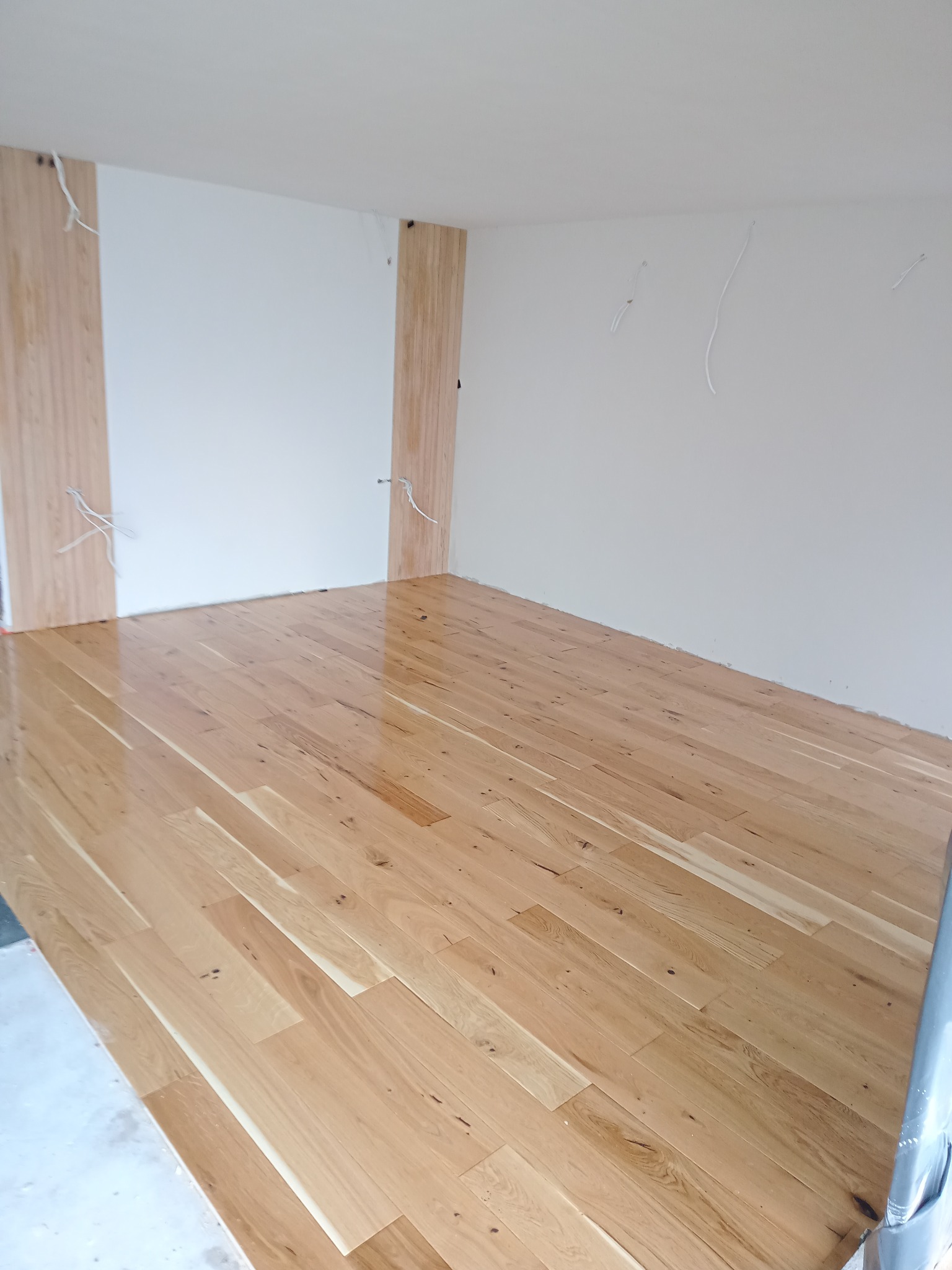 Jasne, lakierowane panele podłogowe w nowym, pustym pokoju z przygotowanymi przewodami elektrycznymi na ścianach.