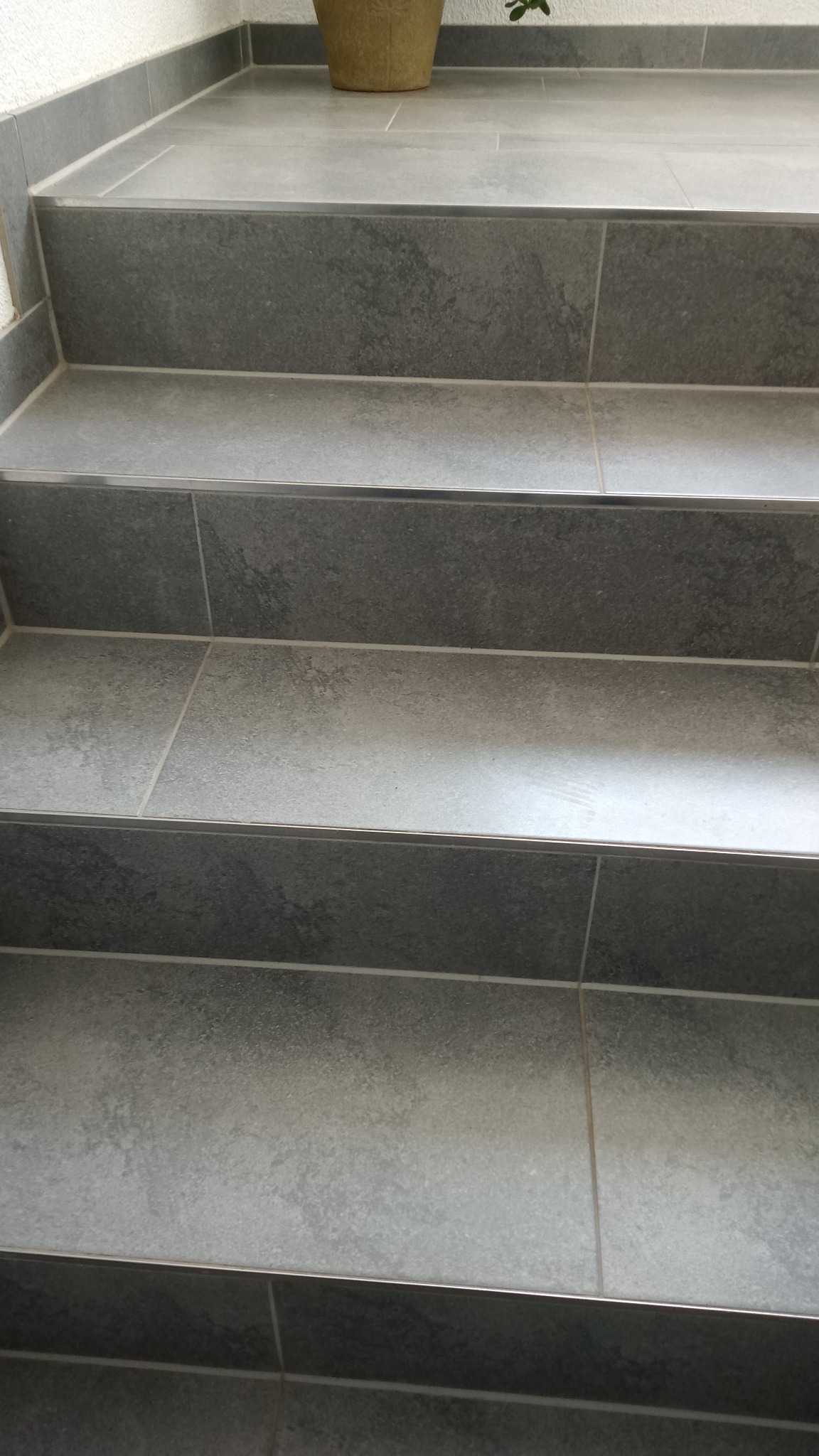 Szare, kamienne schody wyłożone płytkami z metalowymi listwami wykończeniowymi, prowadzące do góry, z doniczką widoczną w tle.