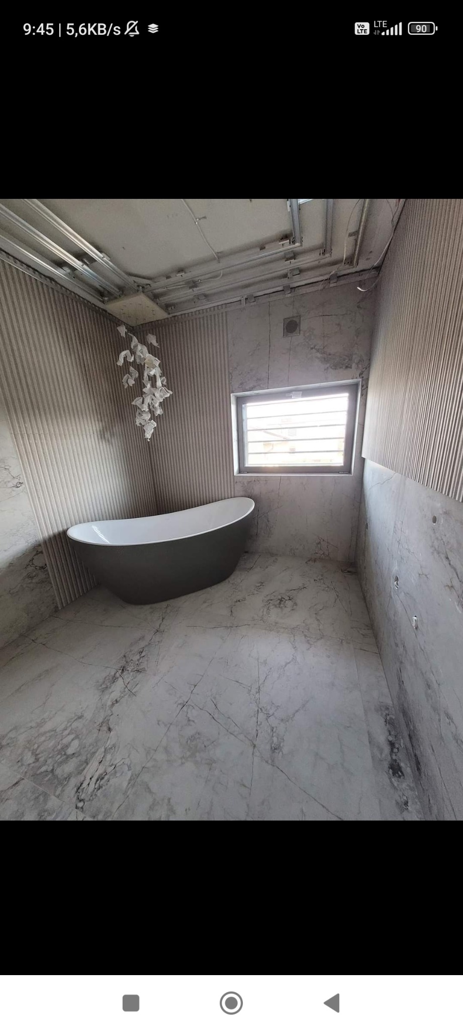 Szara wanna wolnostojąca w trakcie remontu łazienki, widoczne przewody na suficie, częściowo wykończone ściany z pionowymi panelami i imitacją marmuru, podłoga z płytek imitujących marmur.