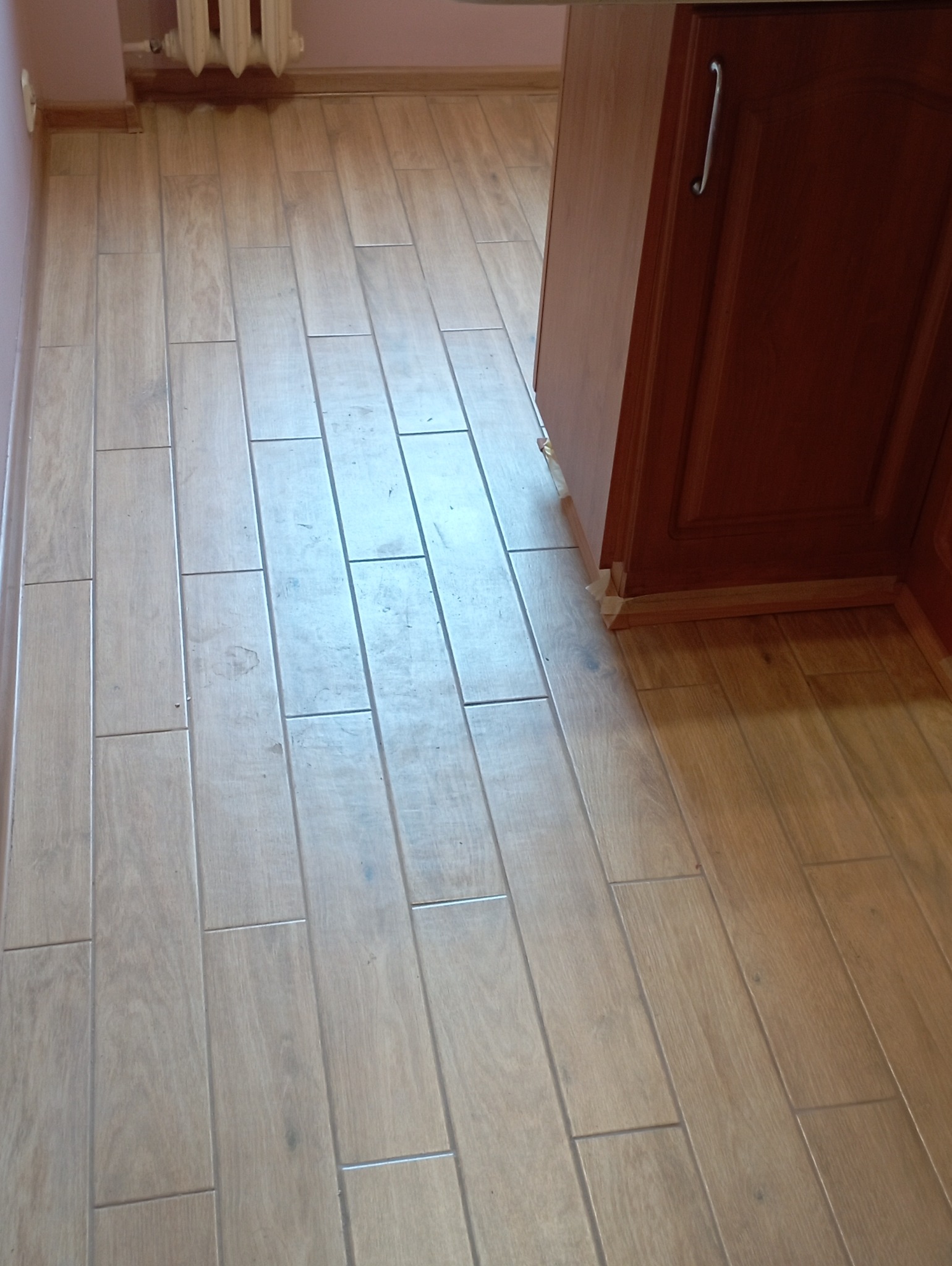 Ułożone podłużne płytki podłogowe imitujące drewno, widoczne przy listwie przypodłogowej i brązowej szafce kuchennej w Wejherowie.