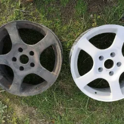 Dwie felgi samochodowe na trawie, jedna szara, brudna, druga pomalowana na biało, ukazujące efekt przed i po renowacji.