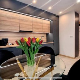 Wykończenia wnętrz Krzyś-Art Krzysztof Włodarczyk - Nowoczesna kuchnia z drewnianymi i grafitowymi szafkami, oświetleniem LED pod szafkami, okrągłym szklanym stołem z bukietem tulipanów i dekoracyjną tapetą z geometrycznym wzorem w odcieniach...