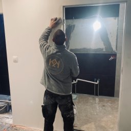 Wykończenia wnętrz Krzyś-Art Krzysztof Włodarczyk - Pracownik w szarej bluzie z logo firmy wygładza ścianę w jasnym pomieszczeniu z widoczną instalacją hydrauliczną w tle