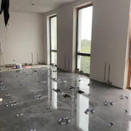 Układanie ciemnych, polerowanych płytek podłogowych z użyciem systemu poziomowania; widoczne okna z widokiem na zieleń i instalacje rur na ścianie.