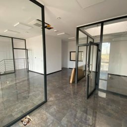 Wykończenia wnętrz Krzyś-Art Krzysztof Włodarczyk - Wnętrze biurowe w trakcie prac wykończeniowych, z widocznymi szklanymi ścianami w czarnych ramach, błyszczącą podłogą imitującą kamień i elementami instalacji wystającymi z posadzki.