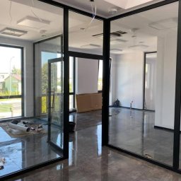 Wykończenia wnętrz Krzyś-Art Krzysztof Włodarczyk - Nowoczesne biuro z przeszklonymi ścianami w czarnych ramach, widoczne materiały budowlane na podłodze, błyszcząca podłoga odbija światło.