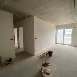 Wykończenia wnętrz Krzyś-Art Krzysztof Włodarczyk - Pokój w stanie deweloperskim po nałożeniu gładzi, widoczne narzędzia i wiaderka z materiałami na podłodze, surowy betonowy sufit z wystającymi kablami, grzejnik pod oknem.