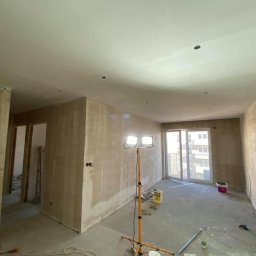 Wykończenia wnętrz Krzyś-Art Krzysztof Włodarczyk - Pokój w trakcie przygotowania do malowania, ściany po szpachlowaniu, widoczne ślady po gładzi, podłoga zabezpieczona, lampa budowlana oświetla wnętrze, okno balkonowe z widokiem na budynek.