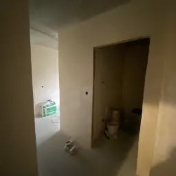 Wnętrze w trakcie remontu: świeżo położone gładzie na ścianach korytarza, worki z gładzią widoczne w oddali, przygotowane puszki elektryczne na podłodze, surowa łazienka w głębi.