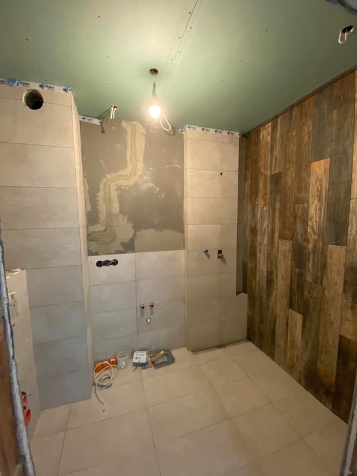 Remontowana łazienka z ułożonymi jasnymi płytkami ściennymi i podłogowymi, fragment ściany wykończony drewnopodobnymi panelami, widoczne instalacje hydrauliczne i elektryczne w trakcie prac.