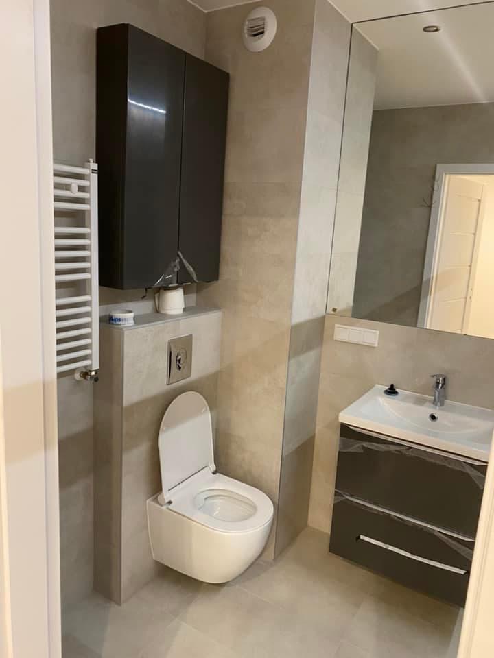 Nowoczesna łazienka w odcieniach szarości z podwieszaną toaletą, grzejnikiem drabinkowym i szafką z umywalką, elementy zabezpieczone folią ochronną.