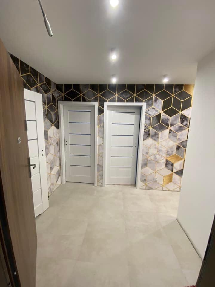 Wykończony korytarz z geometryczną tapetą w odcieniach szarości i złota, dwiema białymi drzwiami z poziomymi frezowaniami i szarą podłogą z płytek w formacie kwadratu, widok z otwartych drzwi.