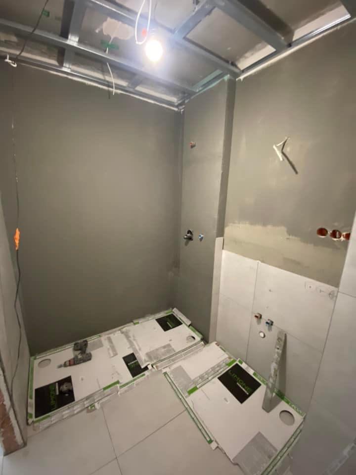 Łazienka w trakcie remontu: ściany po nałożeniu gładzi, częściowo wyłożone płytkami, zabezpieczone podłoże pod płytki na podłodze, widoczne narzędzia (wiertarka) i metalowa konstrukcja pod sufitem.