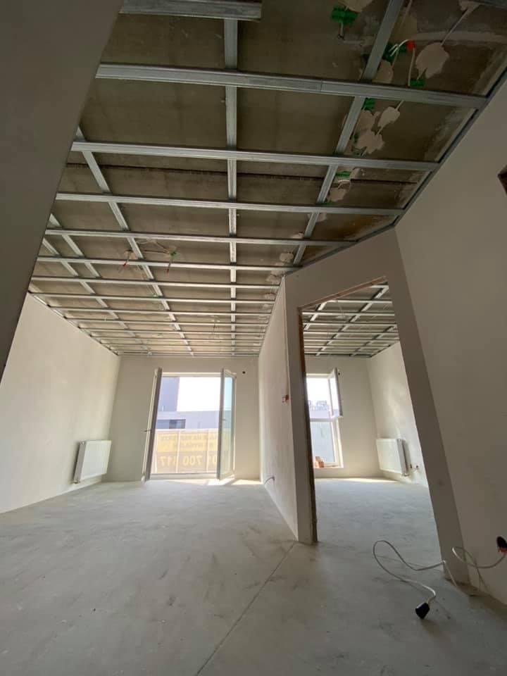 Wnętrze w trakcie montażu sufitu podwieszanego z metalowej konstrukcji, widoczne przewody elektryczne i fragmenty instalacji, ściany przygotowane do dalszych prac wykończeniowych, okna z widokiem...