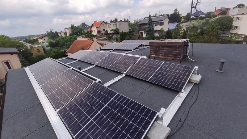 Instalacja paneli słonecznych na płaskim dachu budynku mieszkalnego, widok z góry, z kominem i elementami wentylacji w tle zabudowa jednorodzinna.