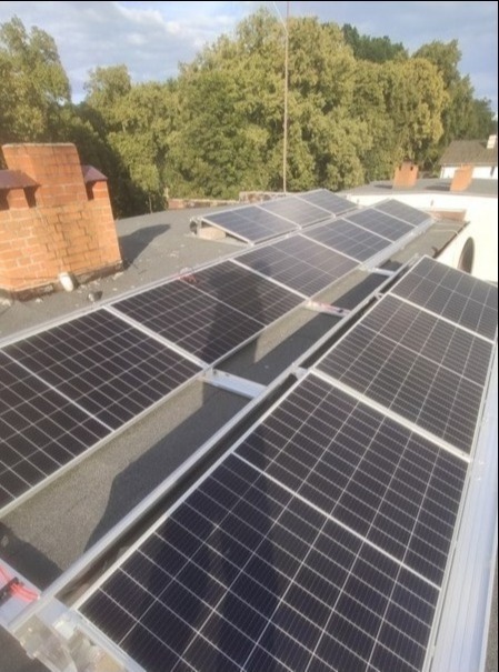 Panele słoneczne zamontowane na płaskim dachu z widocznym kominem z cegły i zielenią drzew w tle.