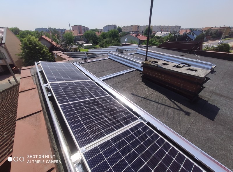 Montaż paneli słonecznych na dachu pokrytym papą, widoczne aluminiowe profile montażowe i ceglany komin w tle zabudowy miejskiej.