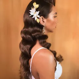 Profil kobiety z ciemnymi, ułożonymi falami włosów, ozdobionych kwiatem i złotymi liśćmi, długie kolczyki, delikatny kontur twarzy, jasna sukienka na tle pomarańczowej ściany.