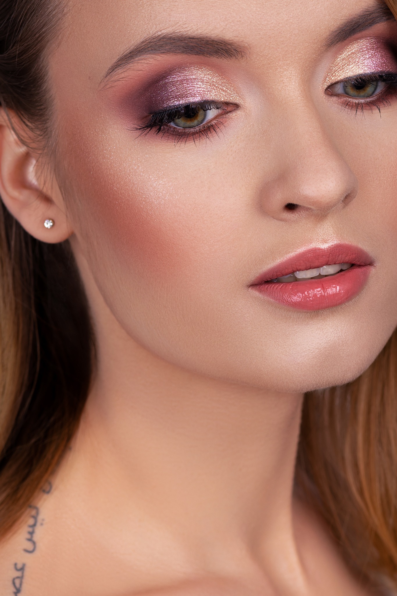 Portret kobiety z profesjonalnym makijażem oczu w odcieniach różu i złota, widoczny kolczyk z brylantem i delikatny makijaż ust w kolorze koralowym, z tatuażem na szyi.
