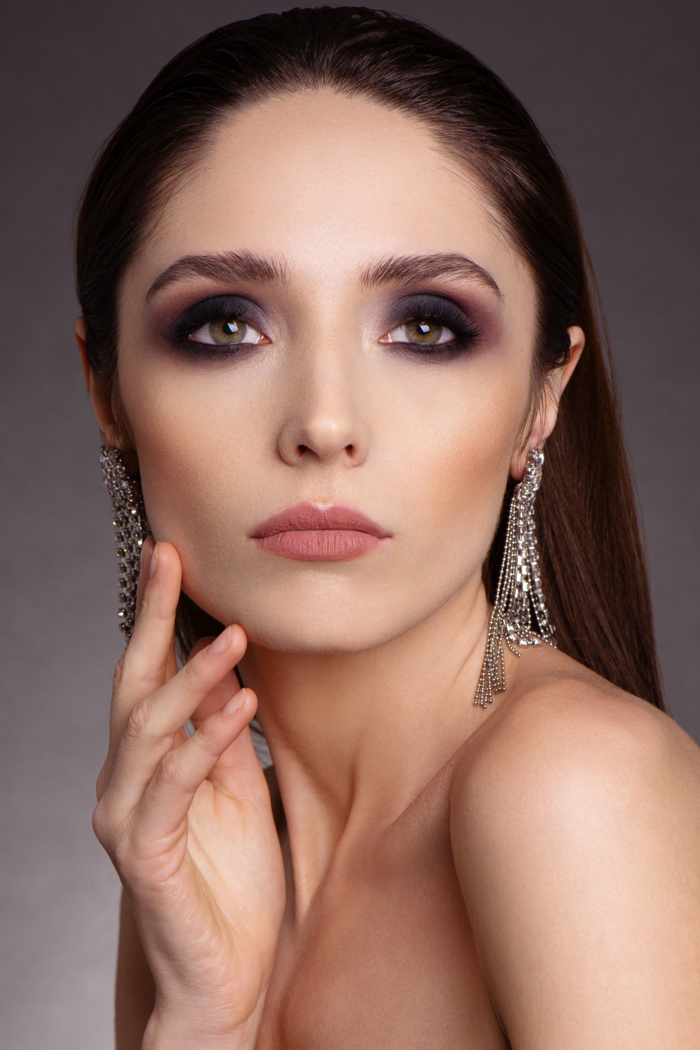 Portret kobiety z intensywnym, przydymionym cieniem na powiekach, podkreślonymi brwiami i matową szminką w odcieniu nude, uzupełniony przez błyszczące, kaskadowe kolczyki.