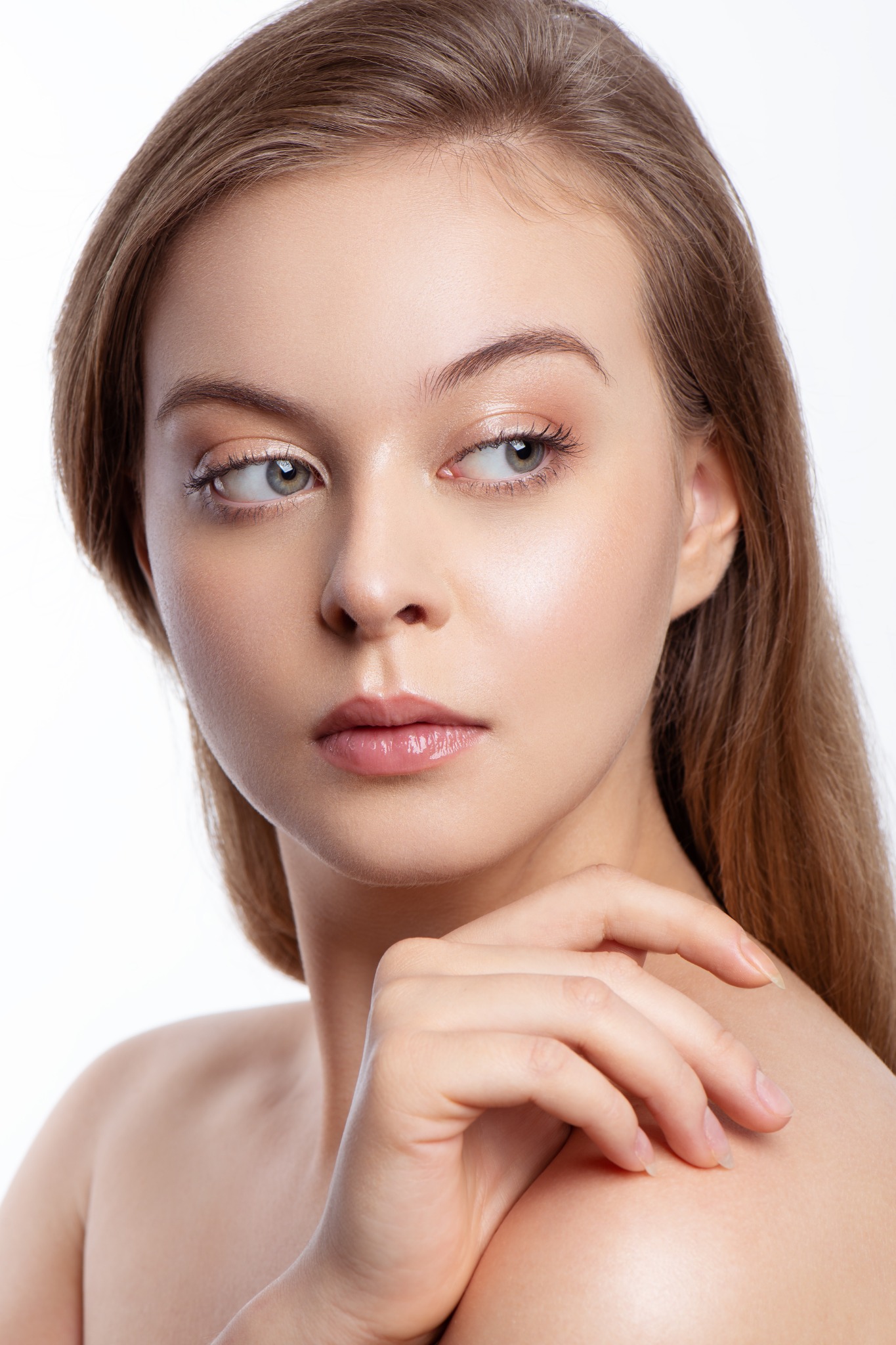 Portret młodej kobiety z subtelnym, naturalnym makijażem, z ręką na ramieniu, spoglądającej w bok na jasnym tle.