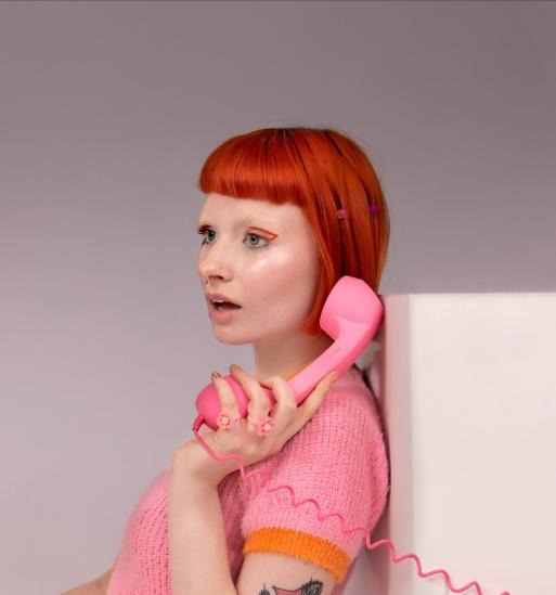 Portret kobiety z rudymi włosami, delikatną kreską na powiekach i różowym telefonem retro, ozdobionym pierścionkami w kształcie kwiatów