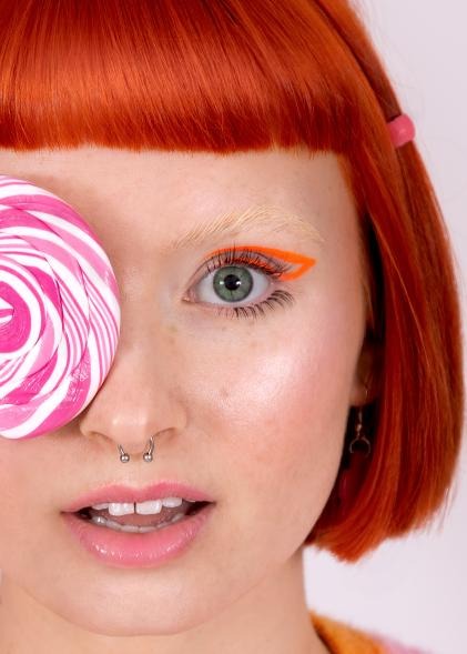 Portret kobiety z pomarańczową kreską na powiece i lizakiem zakrywającym oko, widoczny piercing w nosie i kolczyk w uchu, włosy w odcieniu marchewkowym.
