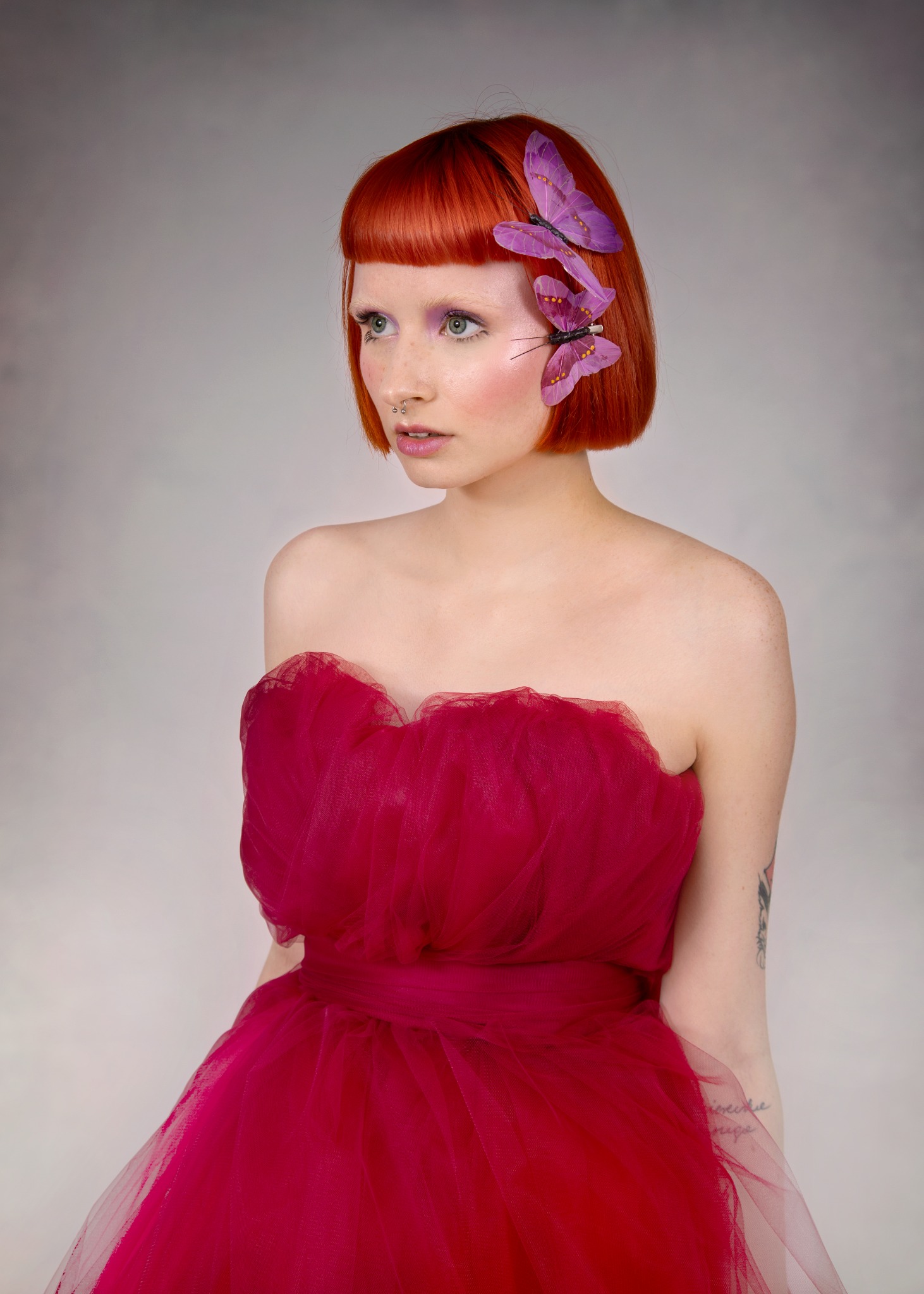 Portret kobiety z rudymi włosami, w różowej sukni, z fioletowymi motylami we włosach, subtelny fioletowy cień na powiekach i delikatny róż na policzkach, piercing w nosie.