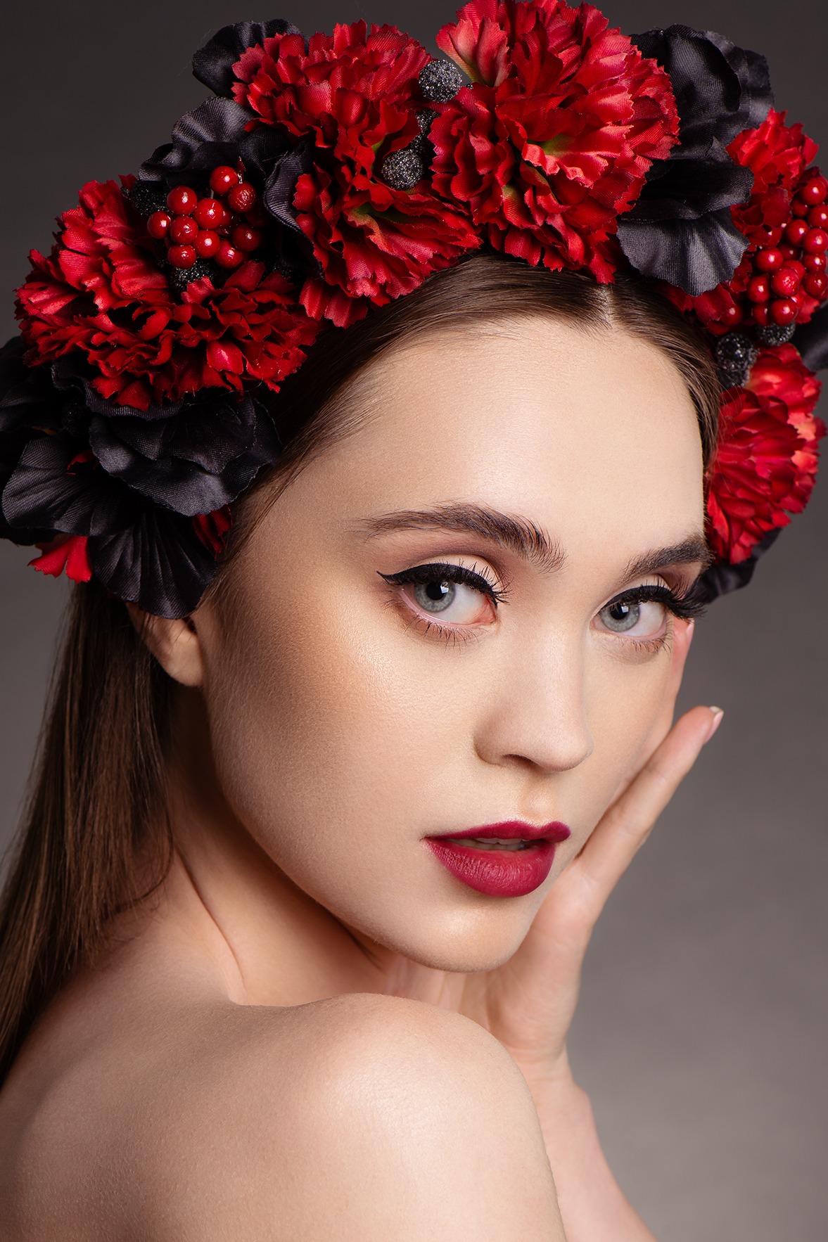 Portret kobiety z delikatnym, wieczorowym makijażem, podkreślonym czarną kreską i ciemnoczerwoną szminką, zwieńczonej wiankiem z czerwonych i czarnych kwiatów, pozującej na szarym tle.