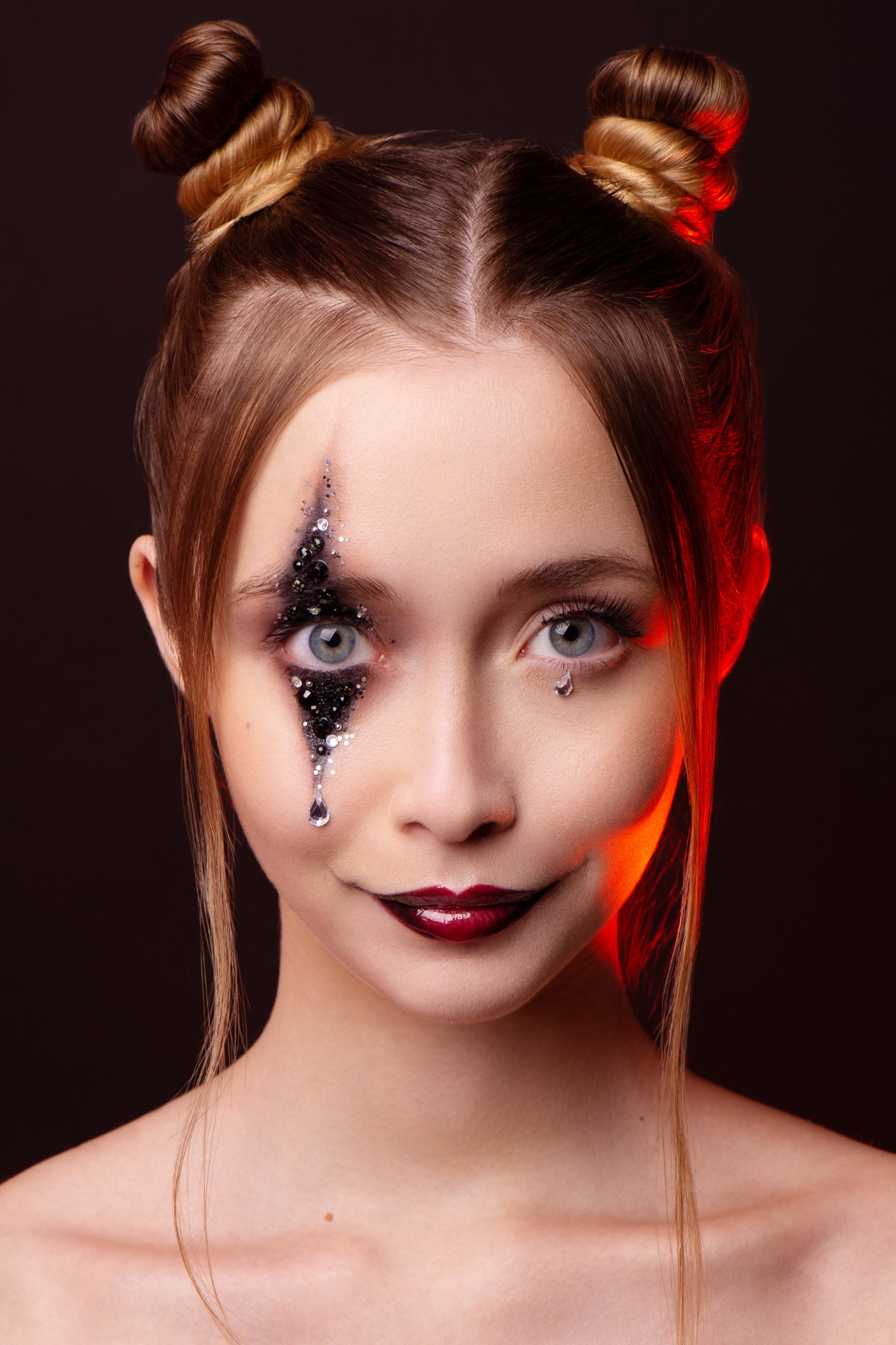 Portret młodej kobiety z artystycznym makijażem: czarne brokatowe zdobienie wokół jednego oka imitujące łzę, ciemna szminka, włosy upięte w dwa koczki. Drugie oko z delikatną, pojedynczą łezką...
