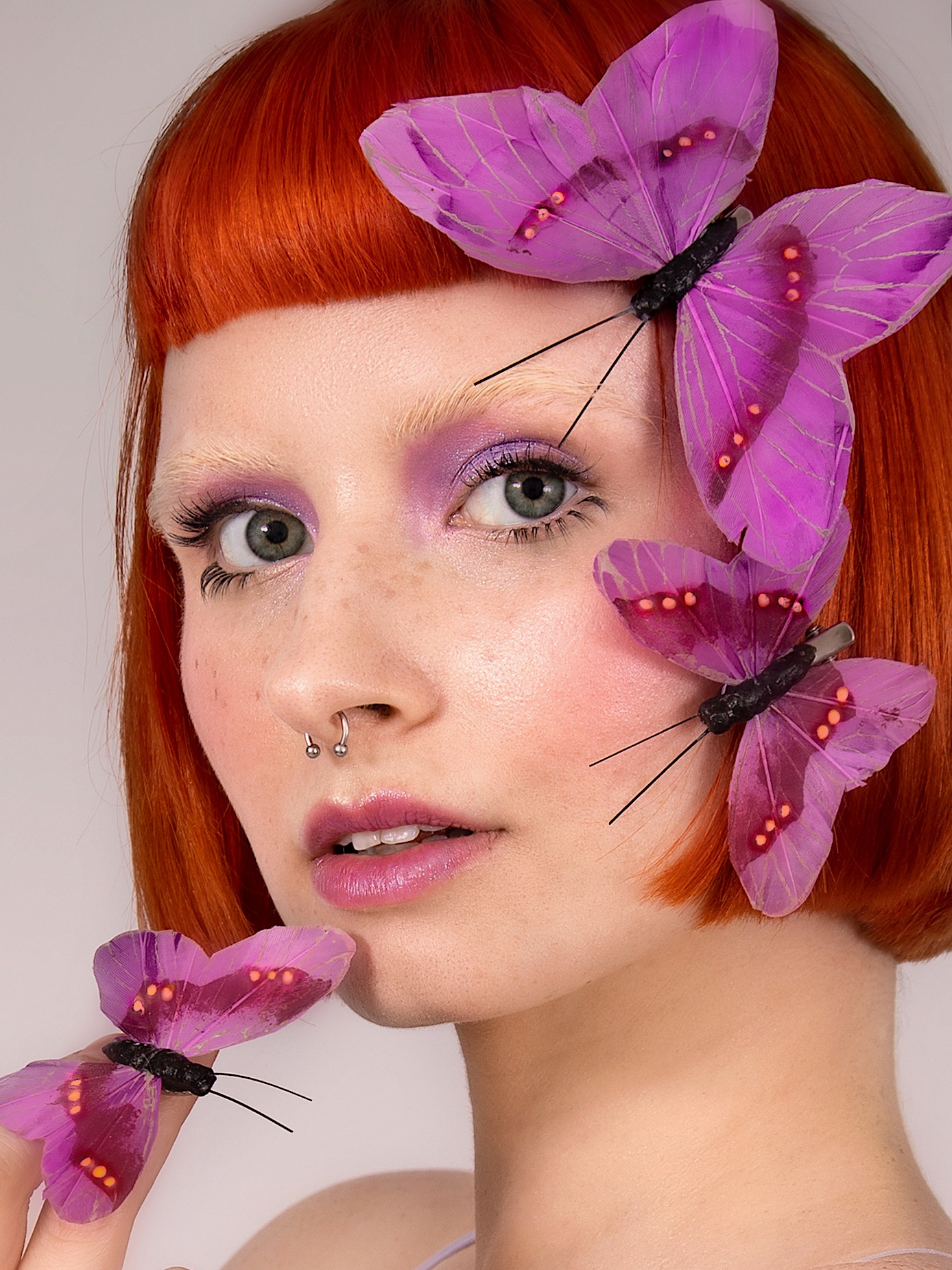 Portret kobiety z rudymi włosami i fioletowym cieniem na powiekach, ozdobiona trzema fioletowymi motylami, z kolczykiem w nosie.