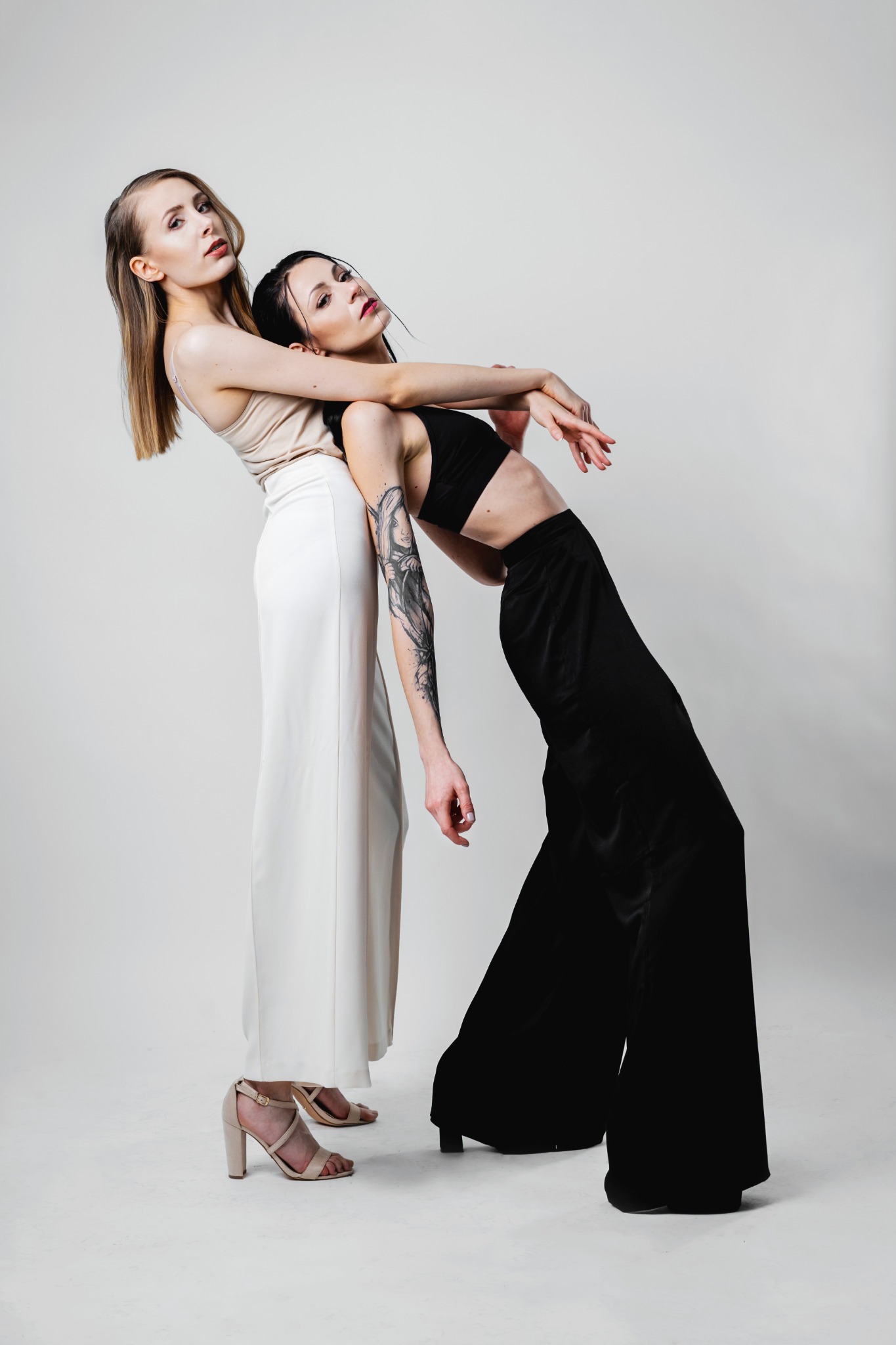 Dwie kobiety w stylizowanych pozach na białym tle: blondynka w beżowej bluzce i białych spodniach opiera się o brunetkę z tatuażem na ramieniu, ubraną w czarny top i szerokie spodnie.