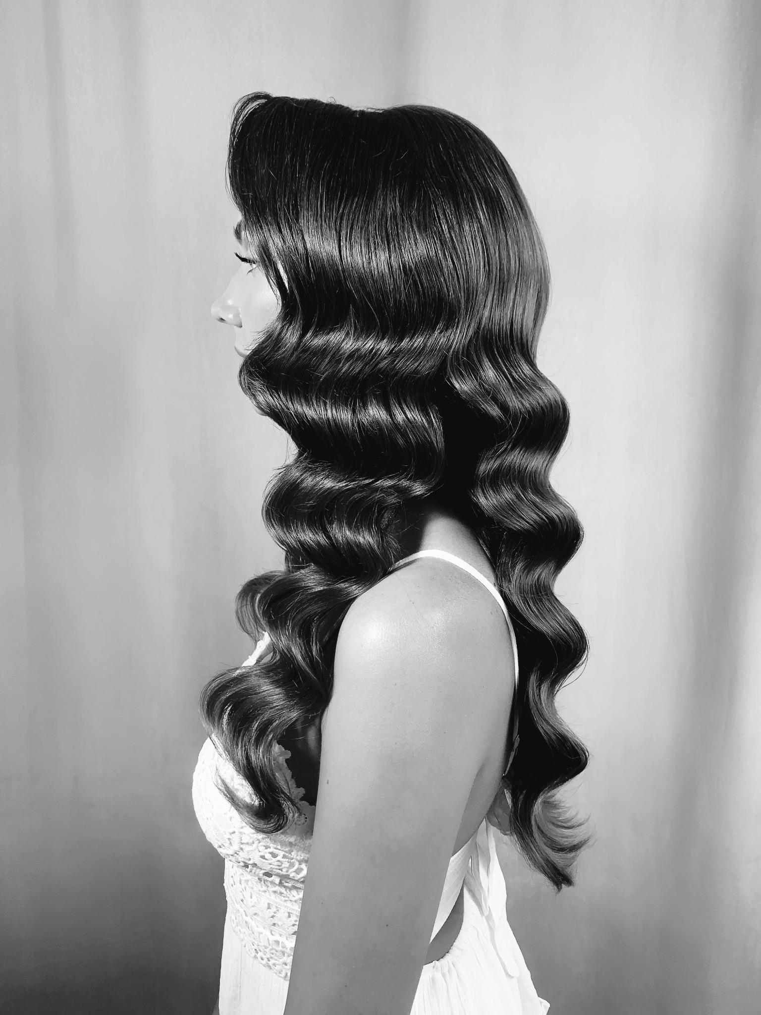 Czarno-białe zdjęcie profilu kobiety z ciemnymi, błyszczącymi włosami ułożonymi w fale w stylu lat 20. i delikatnym makijażem oczu, ubrana w koronkową sukienkę na ramiączkach.