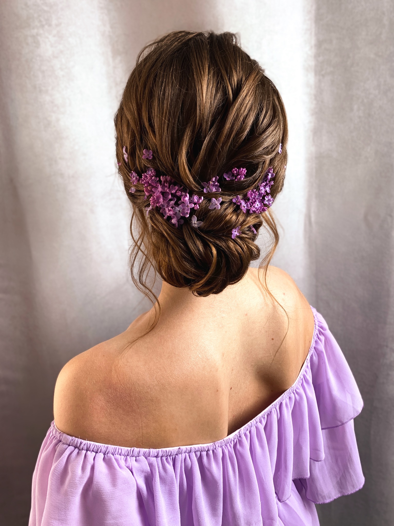 Upięte brązowe włosy z wplecionymi drobnymi fioletowymi kwiatkami, widok z tyłu, odsłonięte ramię kobiety ubranej w liliową bluzkę z falbanami.