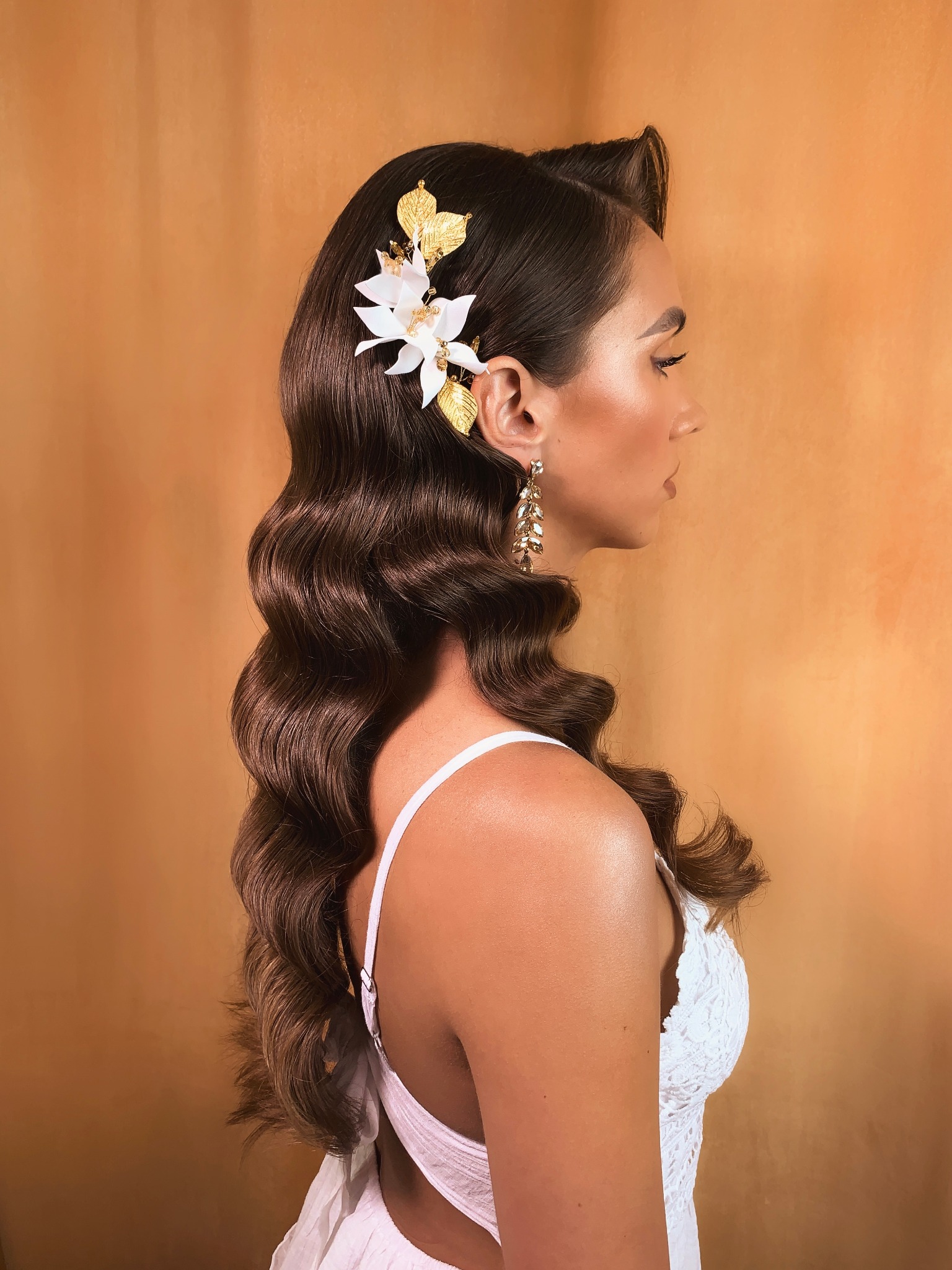 Profil kobiety z ciemnymi, ułożonymi falami włosów, ozdobionych kwiatem i złotymi liśćmi, długie kolczyki, delikatny kontur twarzy, jasna sukienka na tle pomarańczowej ściany.